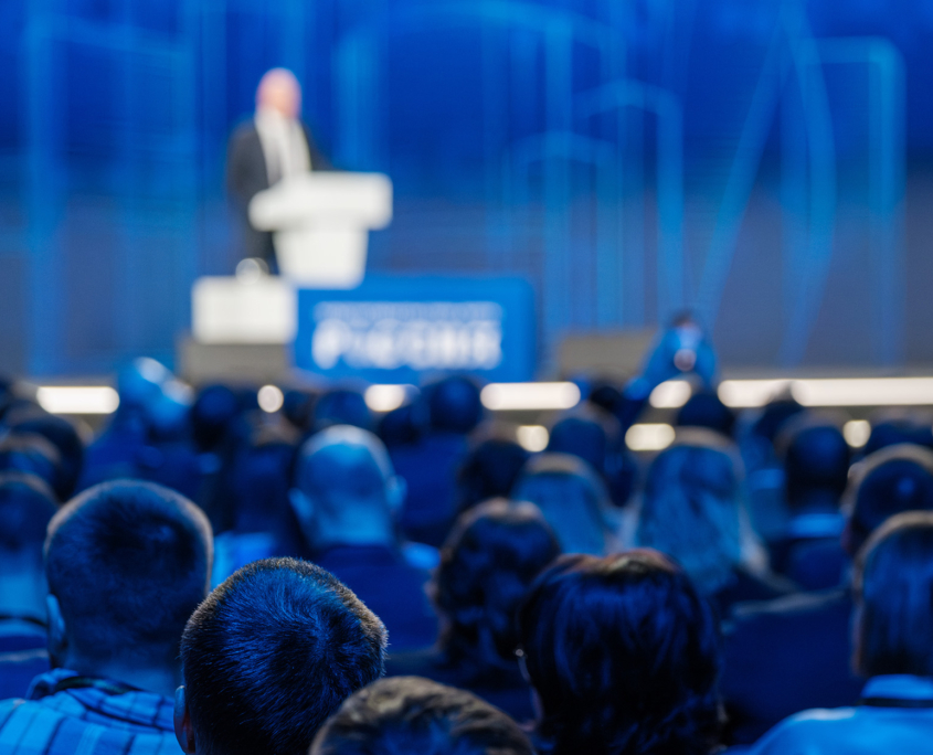 Back View Of Audience Facing Stage Watching Keynote Speaker